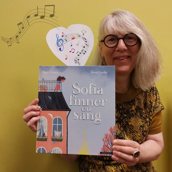 En av bibliotekets ansatte viser frem boka Sofia finner en sang. Hun smiler. Foto. På boka er det en tegning av et hus med vinduer, på en takterasse står en mørkhåret jente. Opp fra boka er det en lapp med tegninger av noter som bølger langs notelinjer. 