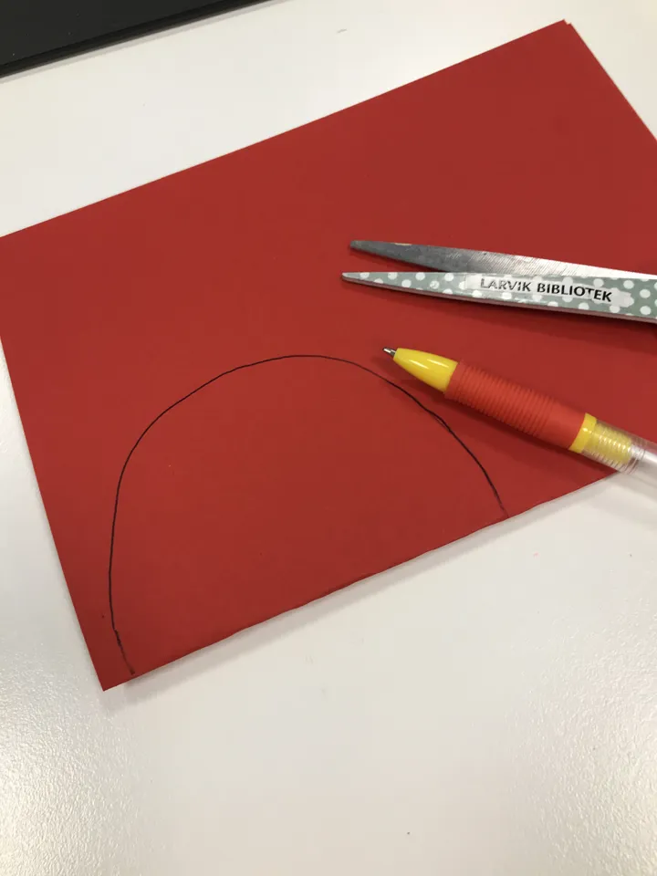 Bildet viser et rødt ark brettet i to, med en tegnet halvsirkel. En penn og ei saks ligger oppå arket. Foto