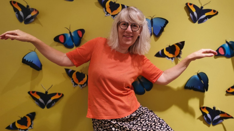 Helga smiler, holder armene ut til siden og står foran en vegg som er fylt med sommerfugler.