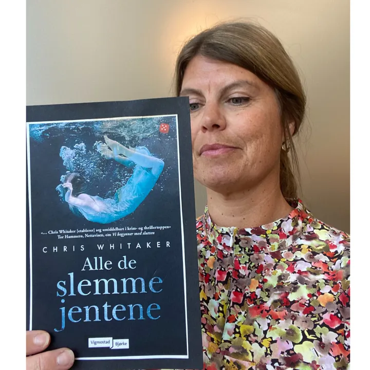 En biblioteksansatt holder boka Alle de slemme jentene opp slik at vi ser omslaget. Hun står og leser bakpå boka , har på seg blomstrete bluse og langt hår i hestehale. Foto. Bokas omslag viser en jente i kjole under vann. Bobler kommer ut fra munnen hennes. På bokas omslag står det med store bokstaver: Chris Whitaker, Alle de slemme jentene.