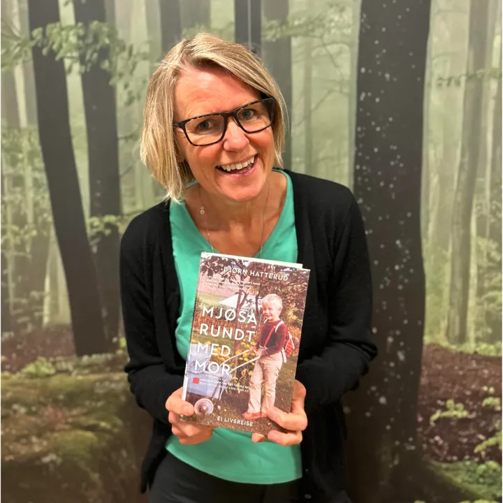 Portrett av ansatt på biblioteket. Hun står foran en vegg med skogmønstrete tapet, smiler og viser frem boka Mjøsa rundt med mor.