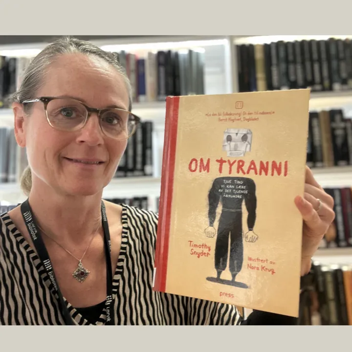 Portrettbilde av en av bibliotekets ansatte som holder boka Om tyranni i hånda si og viser frem forsiden. Hun har briller på og bak henne er en bokhylle med bøker.