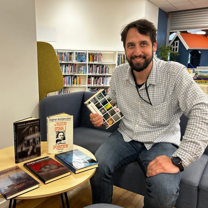 Foto av mannlig ansatt på biblioteket. Han har lys, rutete skjorte, mørkt hår og skjegg og han smiler og sitter i bibliotekets lokaler. I hånden holder han en bok av Ambjørnsen. På et bord ligger flere bøker av Ambjørnsen..