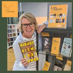 Biblioteksansatt lener seg mot et skap der bøker til bibliotekets litteraturpris står utstilt. Hun smiler og holder frem boka Koke bjørn. Foto.