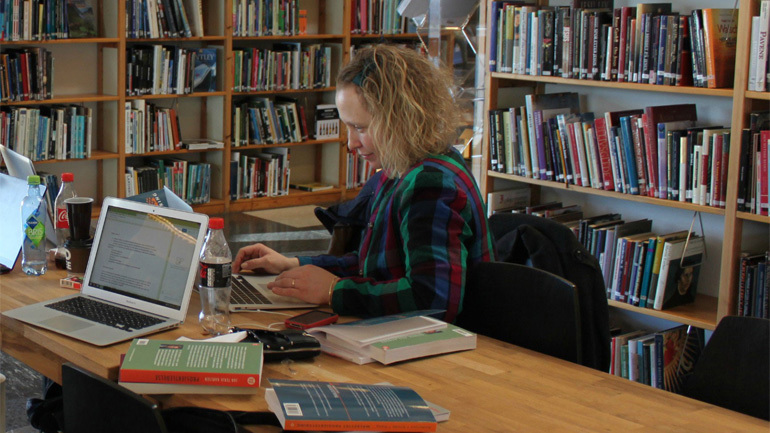 Student sitter i et bibliotek og jobber med en laptop og masse bøker. Foto