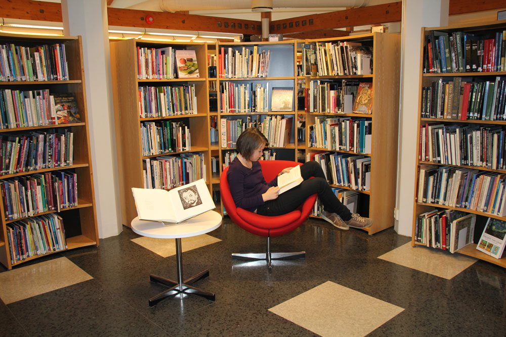 Ung jente leser. Hun sitter i en rød svingstol, bena over armlene. Bokhylle i bue bak. Foto