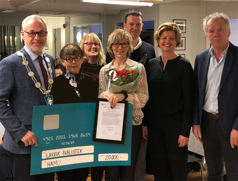 Ordfører og biblioteksjef pluss fem andre voksne personer. Biblioteksjefen holder et bilde (plakett) og roser. Foto