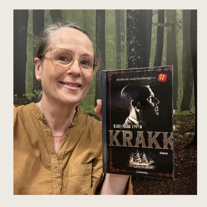 Foto av bibliotekets ansatte. Hun smiler, har på brun skjorte, briller og håret bort fra ansiktet. I den ene hånden viser hun fram forsiden på boka Krakk.
