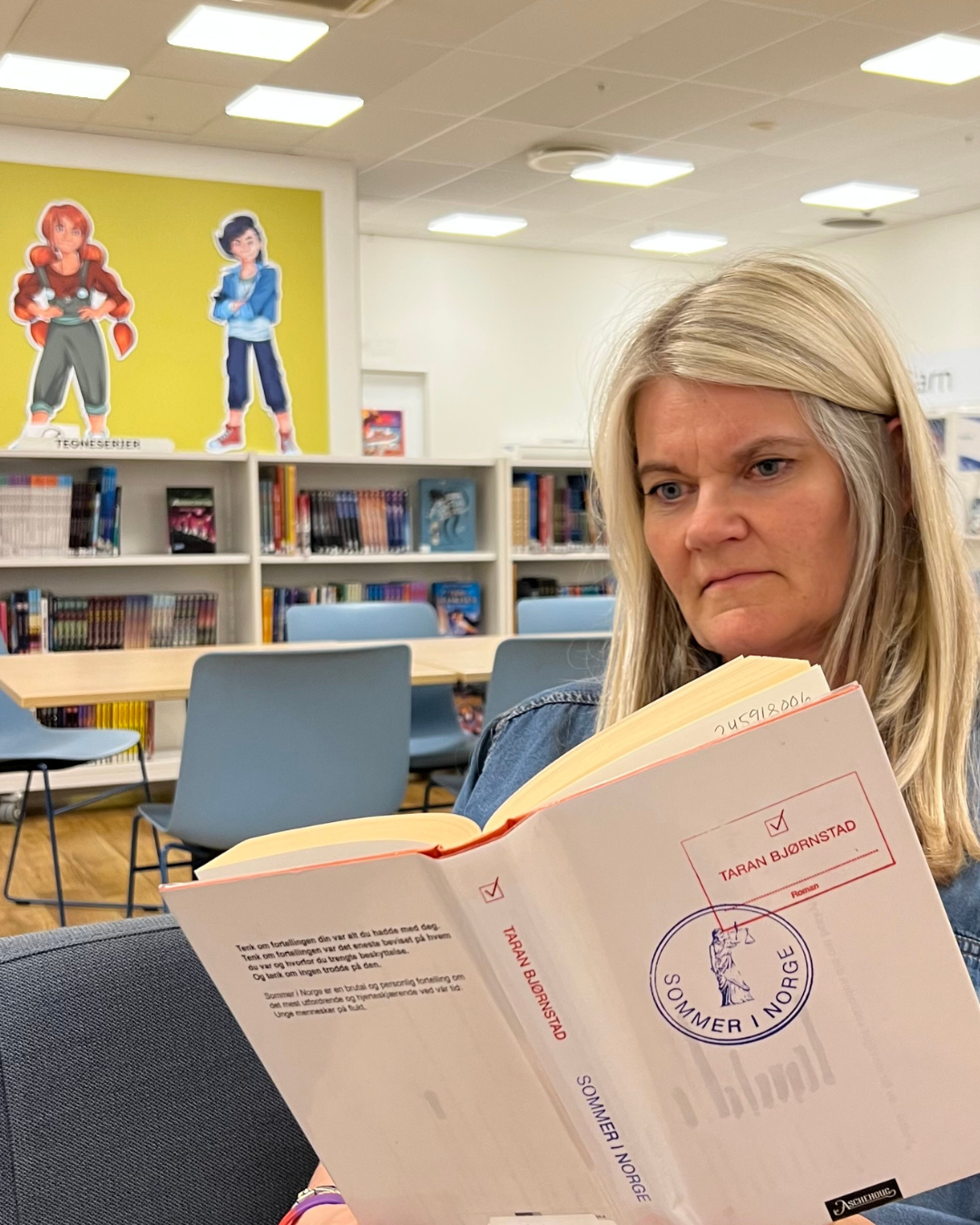 En lyshåret kvinne sitter i et biblioteksrom og leser. Bak henne er det bokhyller med bøker, et langbord med stoler. Foto.