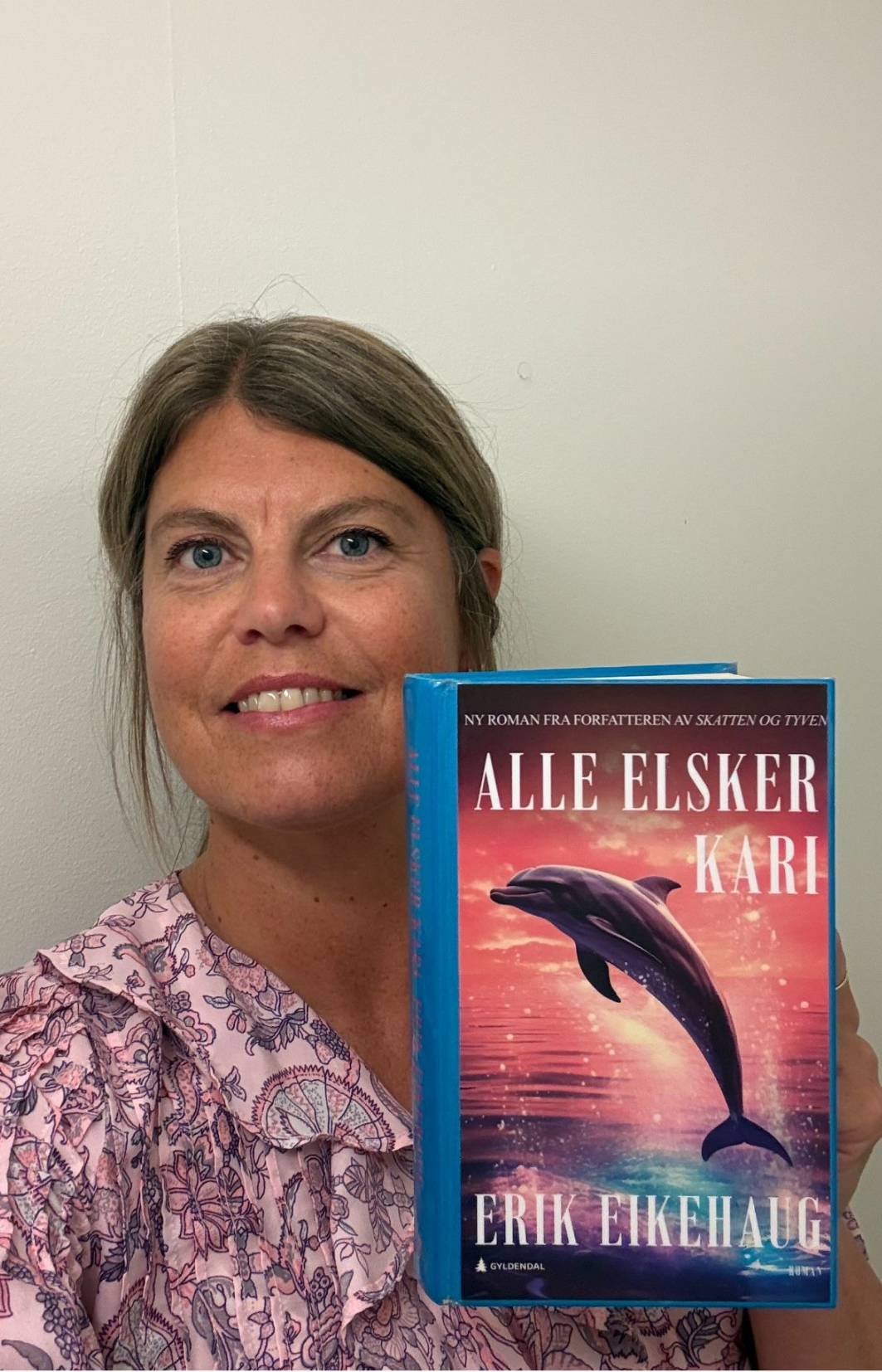 Nærbilde av en dame som smiler og håret satt bort fra ansiktet. Hun har mønstrete bluse og holder frem boka Alle elsker Kari. Foto.