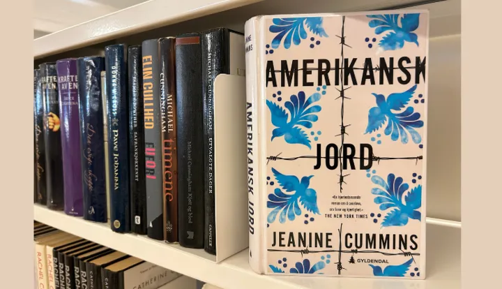 Boka står i en bokhylle og bokas forside vises. Foto. På bokomslaget: Skrift som står på omslaget på boka er: Amerikansk jord står med stor skrift. I tillegg står forfatterens navn og et sitat fra The New York Times som sier: "En hjerteskjærende roman om å overleve, om farer og kjærlighet". Bak skriften er det tegnet piggtråder og fugler som flyr.