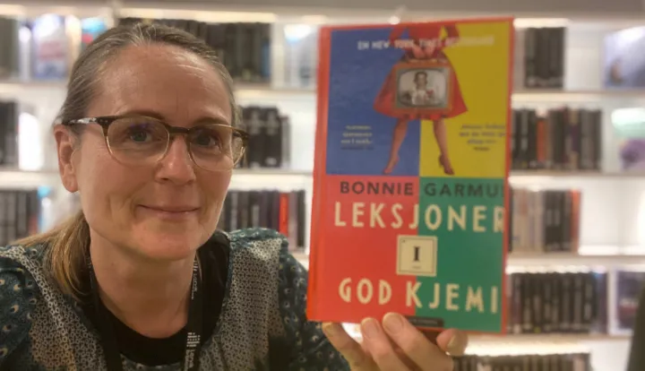 Nærbilde av en av bibliotekets ansatte som holder frem boka Leksjoner i god kjemi. Hun ser inn i kameraet, smiler og har briller på. Foto. På bokomslaget er det en collage med skarpe farger, bokas tittel og forfatterens navn står med store bokstavtyper og over skriften er det tegning av en dame i vidt skjørt med forkle over skjørtet.