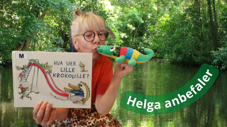 Helga holder boka Hva sier Lille Krokodille i den ene hånden og en tøykrokodille i den andre hånden. Bak henne er det en jungel med grønne trær og vann.