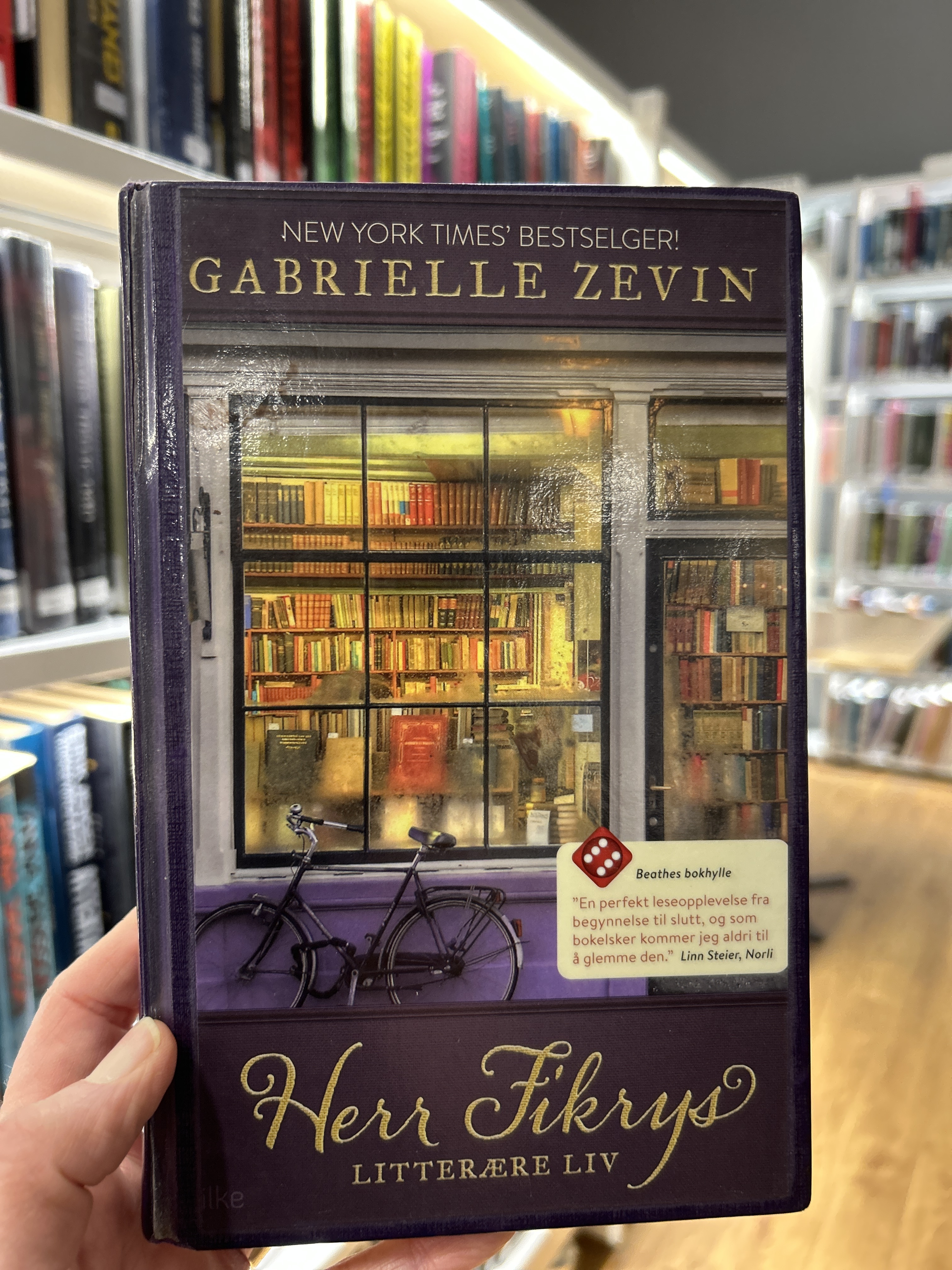 Bokomslag  til boka Herr Fikrys litterære liv. Bildet på boka viser et vindu inn til en bokhandel med hyller fulle av bøker.  Det er lys i butikken. På utsiden av vinduet står en sykkel. 