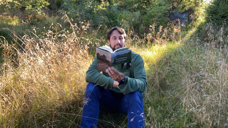 Kjetil sitter i en eng og leser i boka Livet på landet.