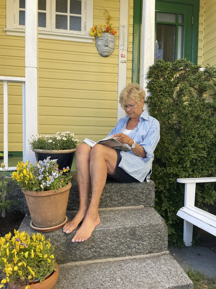 En kvinne sitter på trappen til et gult hus. Det er blomster på trappetrinnene. Hun leser i en bok. Foto: privat