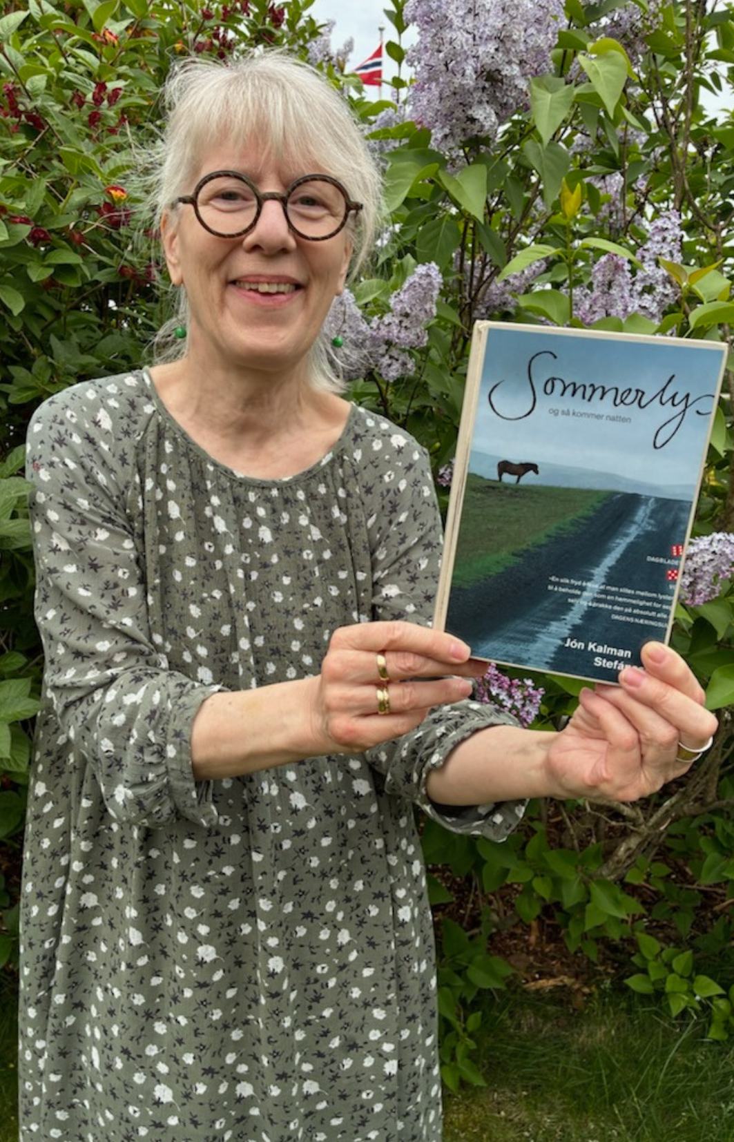 En dame som smiler står foran en blomstrende syrinbusk. Hun har briller, lyst hår og smiler. I hendene holder hun boka Sommerlys-og så kom natten. Foto.