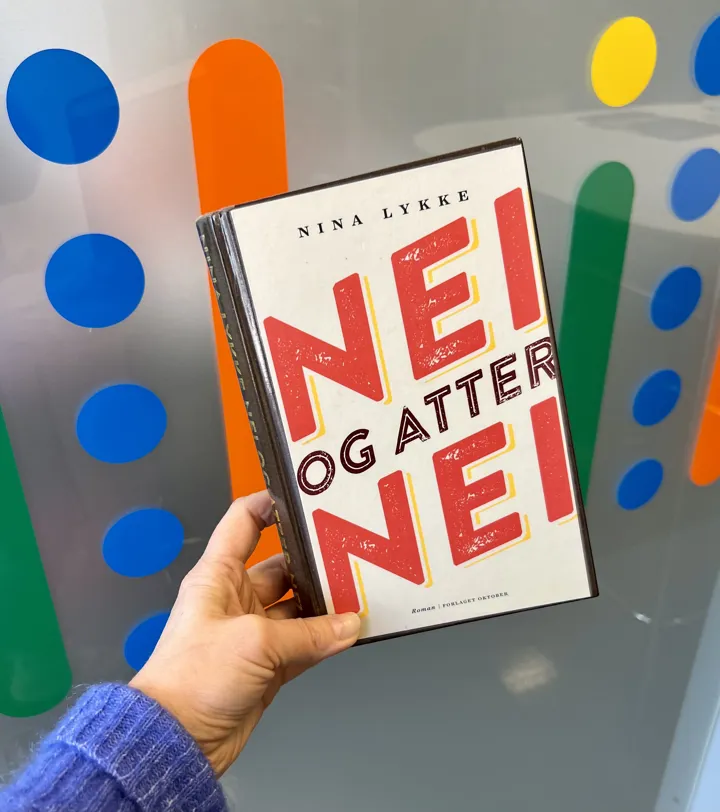 Nærbilde av en hånd som holder boka Nei og atter nei av Nina Lykke. Bak boka er det detaljer fra en glassvegg med fargerike prikker og streker. Tekst som fyller bokomslaget: Nei og atter nei.