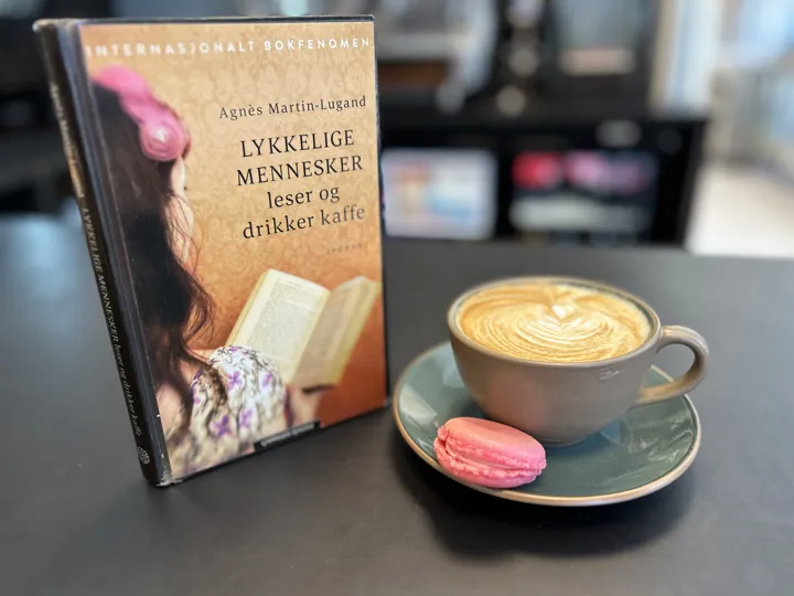 Boka "Lykkelige mennesker..." står på disken til Bokbiten sammen med en grønn kopp med cappuccino og en knall rosa macron liggende på kaffeskåla. Foto