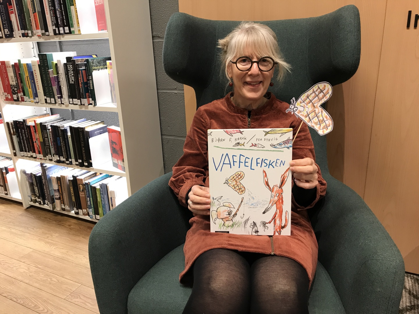 Helga holder en bok som heter vaffelfisken. Hun sitter i en god lenestol og ser inn i kamera. Foto