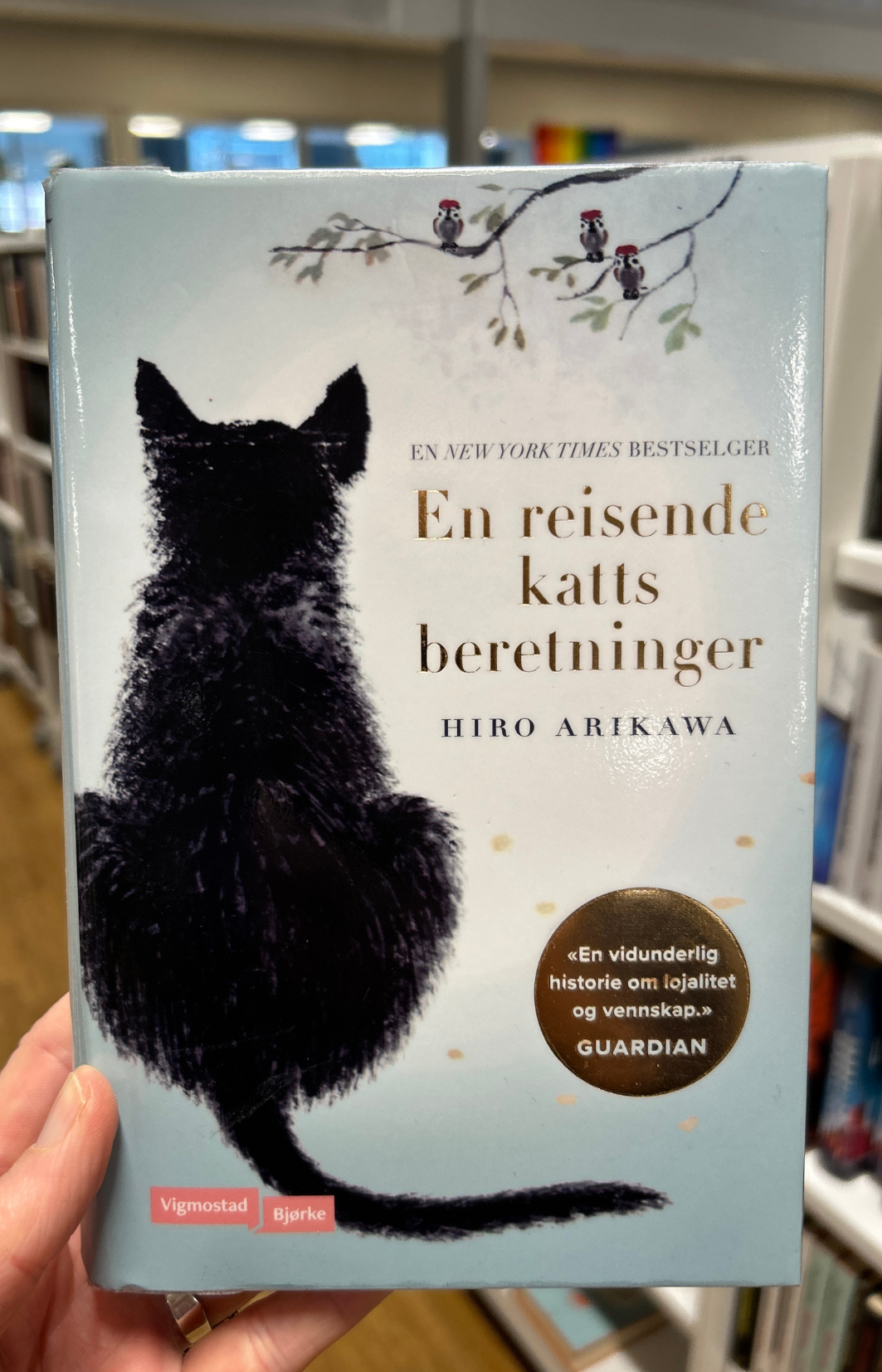Foto av en hånd som holder en boka En reisende katts beretninger. Bak boka sees noen bokhyller med bøker. Bokomslaget viser en tegning av ryggen på en katt som sitter. Over katten er en grein med blader og småfugler på. Tittelen og forfatterens navn står ved siden av katten. til boka 