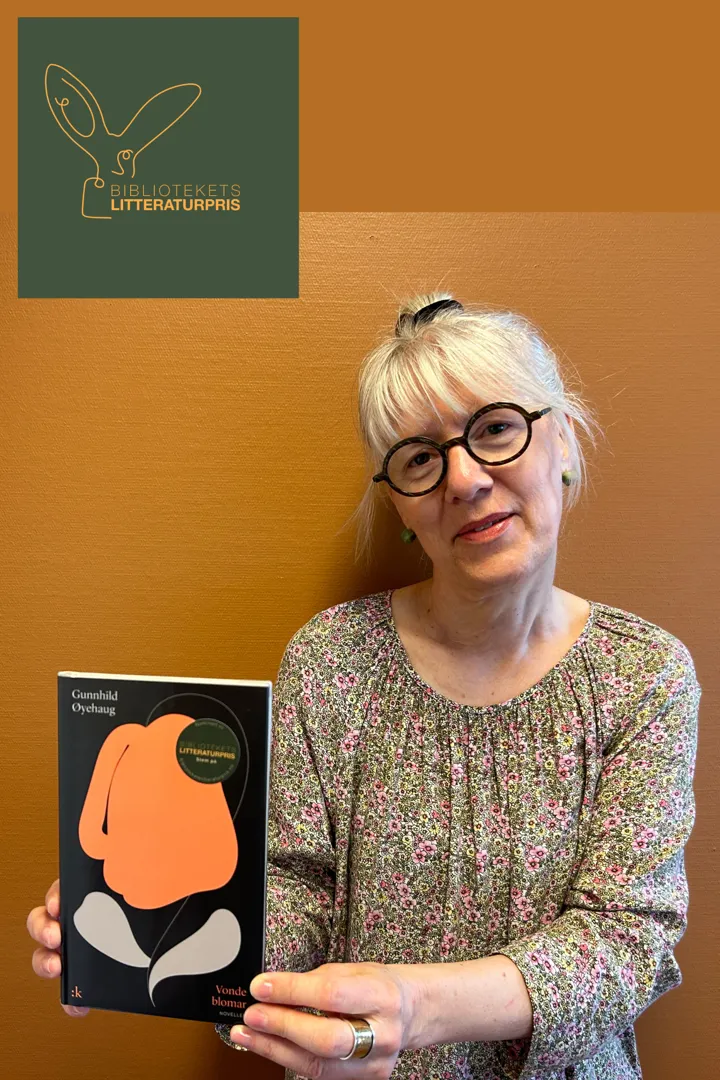 Portrettbilde av biblioteksansatte Helga. Hun står foran en vegg, har småblomstrete kjole, briller og smiler mens hun viser frem boka Vonde blomar.