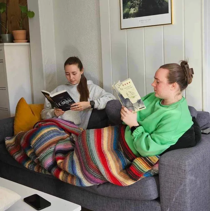 To unge jenter sitter sammen i en sofa. De leser i hver sin bok, og har et fargerikt teppe over beina (de deler dette teppet mellom seg). Foto