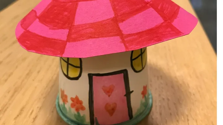 Et søtt lite fe-hus laget av pappbeger og papir. Taket er rutete i to rosa farger, døra er rosa, det er blomster nederst og vinduene er gule. Foto