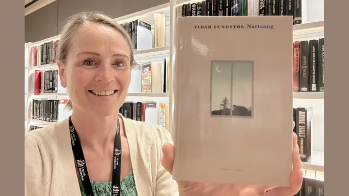 portrettbilde av en av bibliotekets ansatte, hun smiler og ser inn i kameraet og i hånden holder hun frem og viser frem boka Nattsang sin forside. Bak henne er det bokhyller med bøker.Foto. På bokas omslag er det en grafisk fremstilling av et vindu. Utenfor vinduet er det en halvmåne og noen mørke trær.
