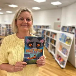 Biblioteksansatt står foran bokhylle i barneavdelingen og i hendene holder hun frem boka Revefarmen. Foto.