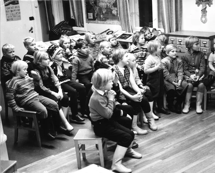 Mange barn sitter på små krakker og lytter. Bildet er i svart hvitt. Foto