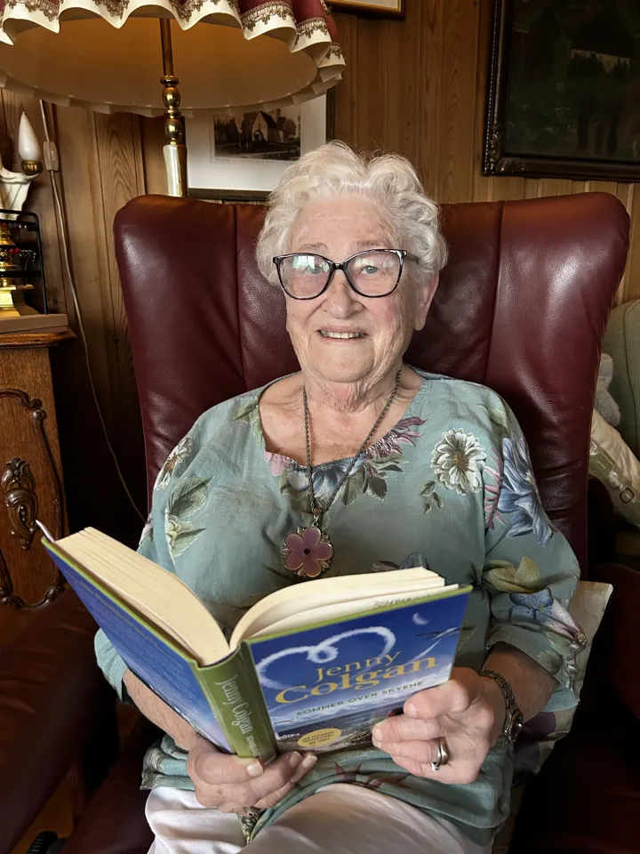 En eldre kvinne sitter en godstol (skinnstol med armlener) og leser i en bok. Hun smiler til kamera. Foto