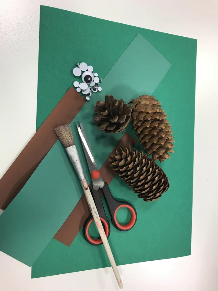 Bildet viser saks, kongler, ferdige øyne til å klistre på, papir i brunt og grønt, en pensel. Foto